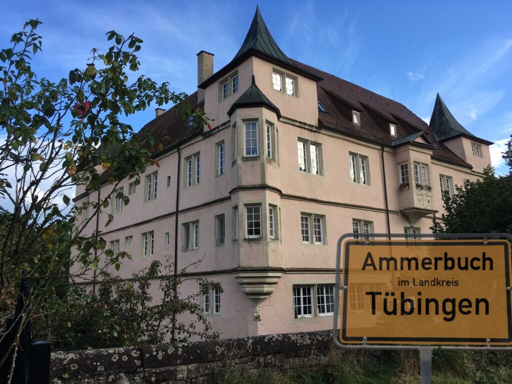 A pink historic building with turreted rooflines beside a stone wall, with a yellow town sign reading 'Ammerbuch im Landkreis Tübingen' in the foreground.