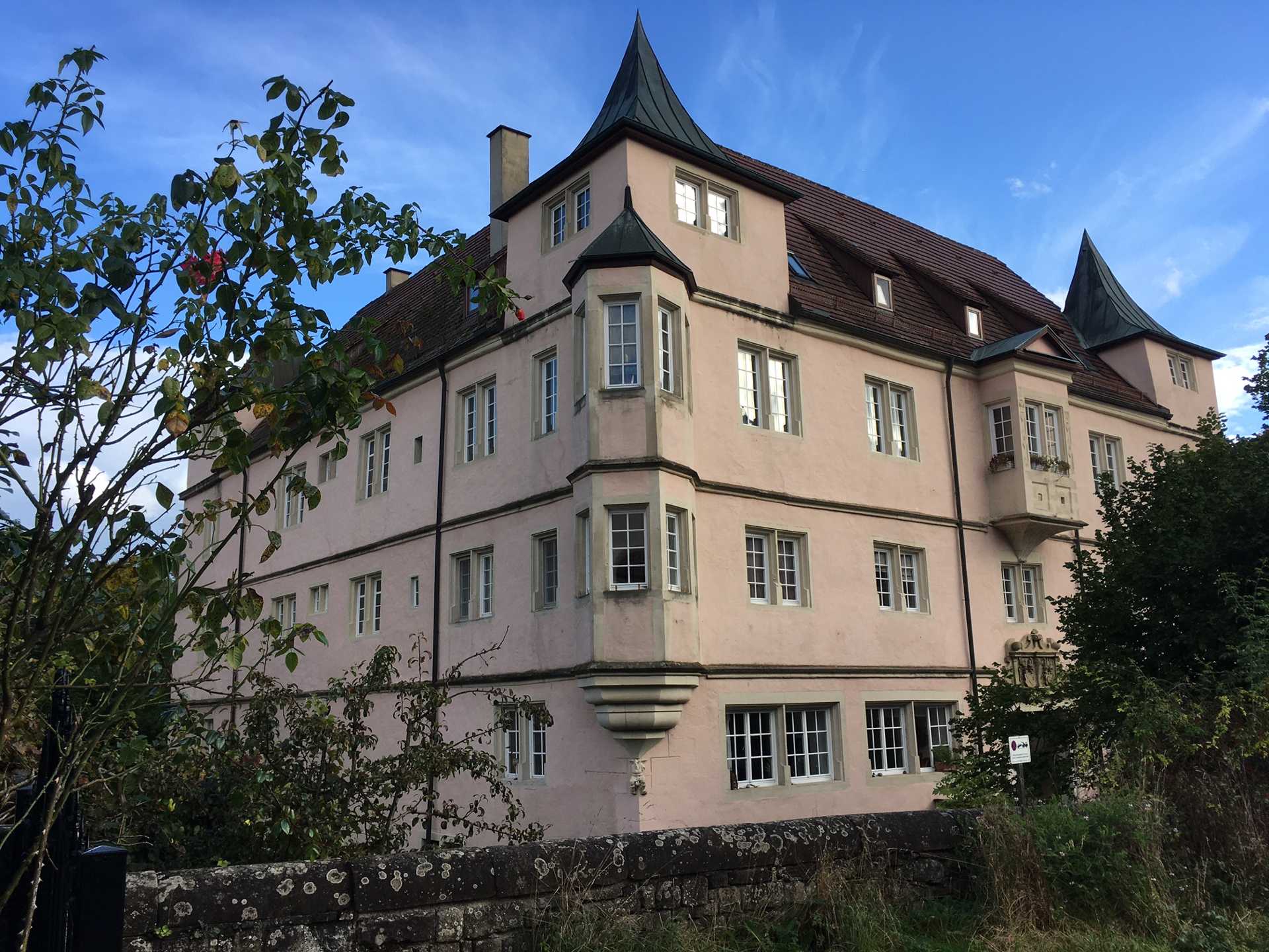 Pink, manor-style building with tower-like roofs, set against a blue sky; stone base and many white-framed windows surrounded by greenery
