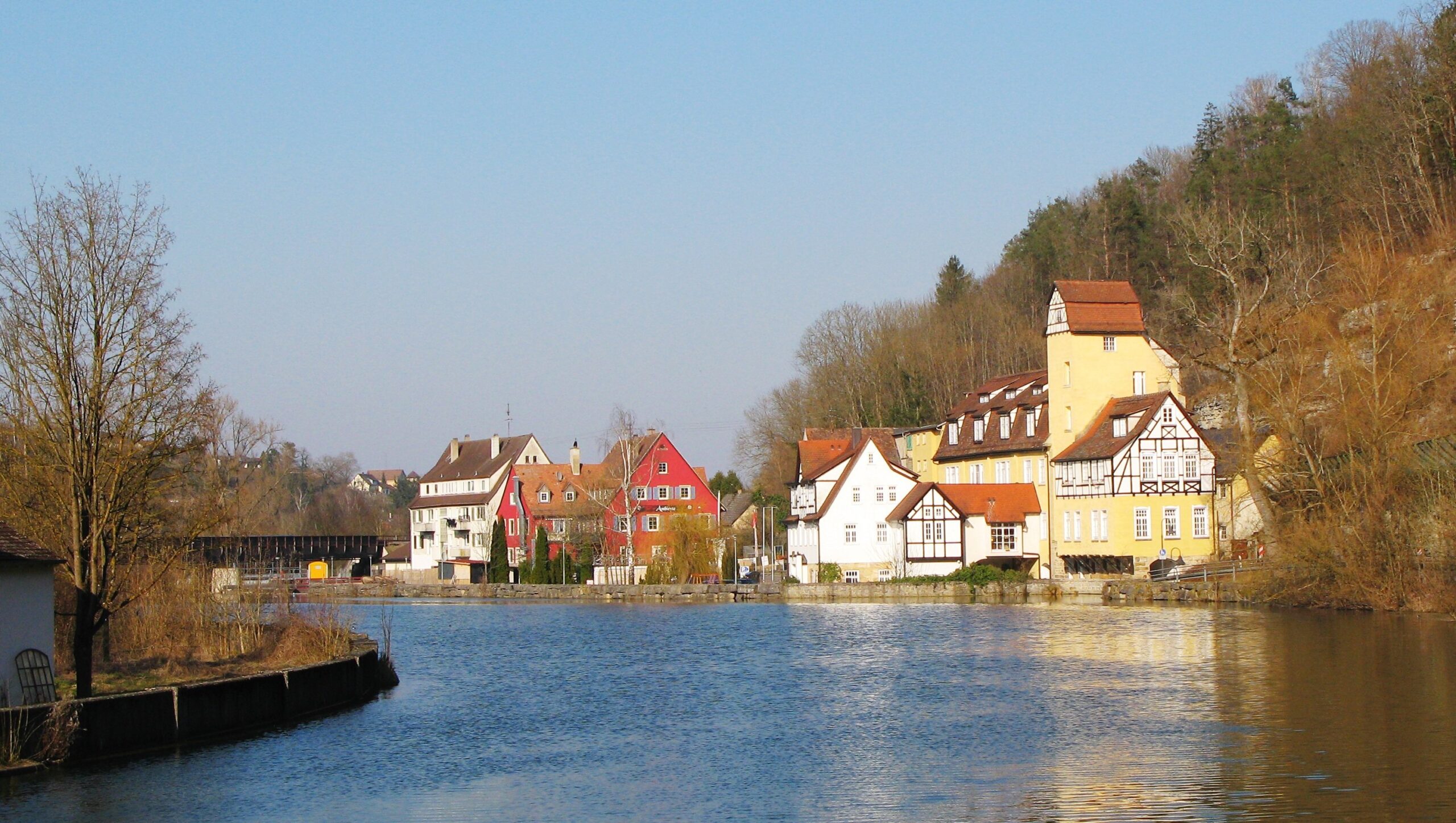 Colorful houses along a calm river with a tree-covered hill in the background.",